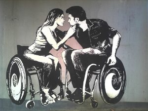 Painted graffiti shows a man and a woman leaning in to kiss each other. They are both wheelchair users
