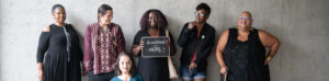 Six disabled people of color smile and pose in front of a concrete wall. Five people stand in the back, with the Black woman in the center holding up a chalkboard sign reading "disabled and here." A South Asian person in a wheelchair sits in front.