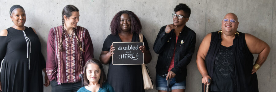 Six disabled people of color smile and pose in front of a concrete wall. Five people stand in the back, with the Black woman in the center holding up a chalkboard sign reading "disabled and here." A South Asian person in a wheelchair sits in front.